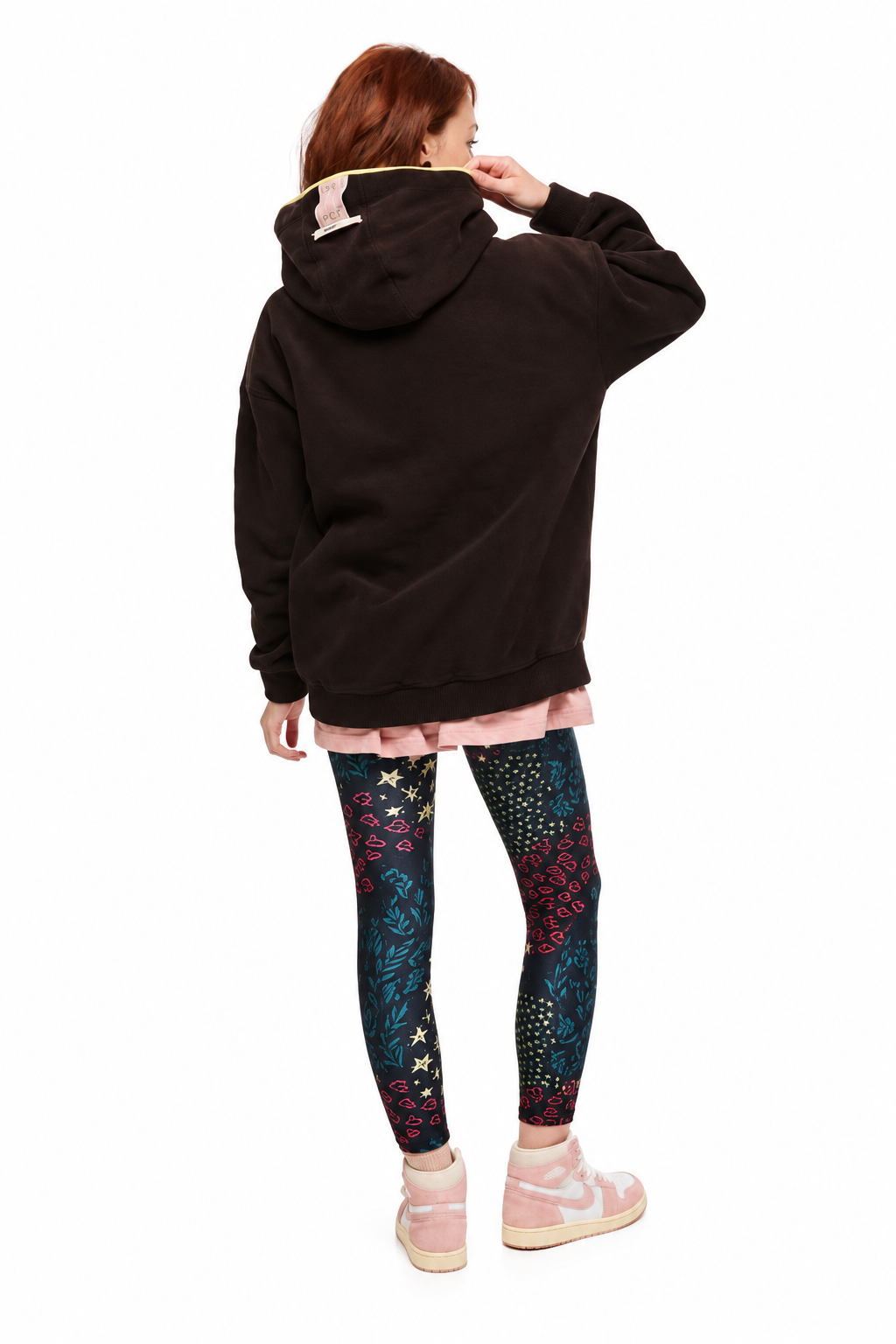 A model shown from the back wearing a black oversized hoodie with a pink bunny ear detail on the hood, a light pink pleated skirt, patterned leggings, and pink sneakers, set against a plain white background.