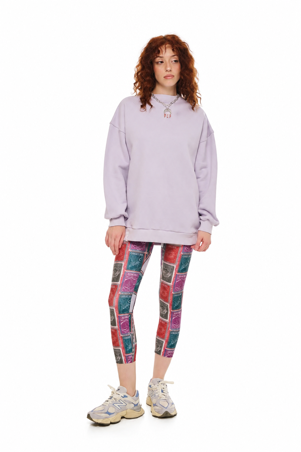 A model stands against a white studio background wearing a light lavender oversized sweatshirt, colorful patchwork print leggings, and white sneakers.