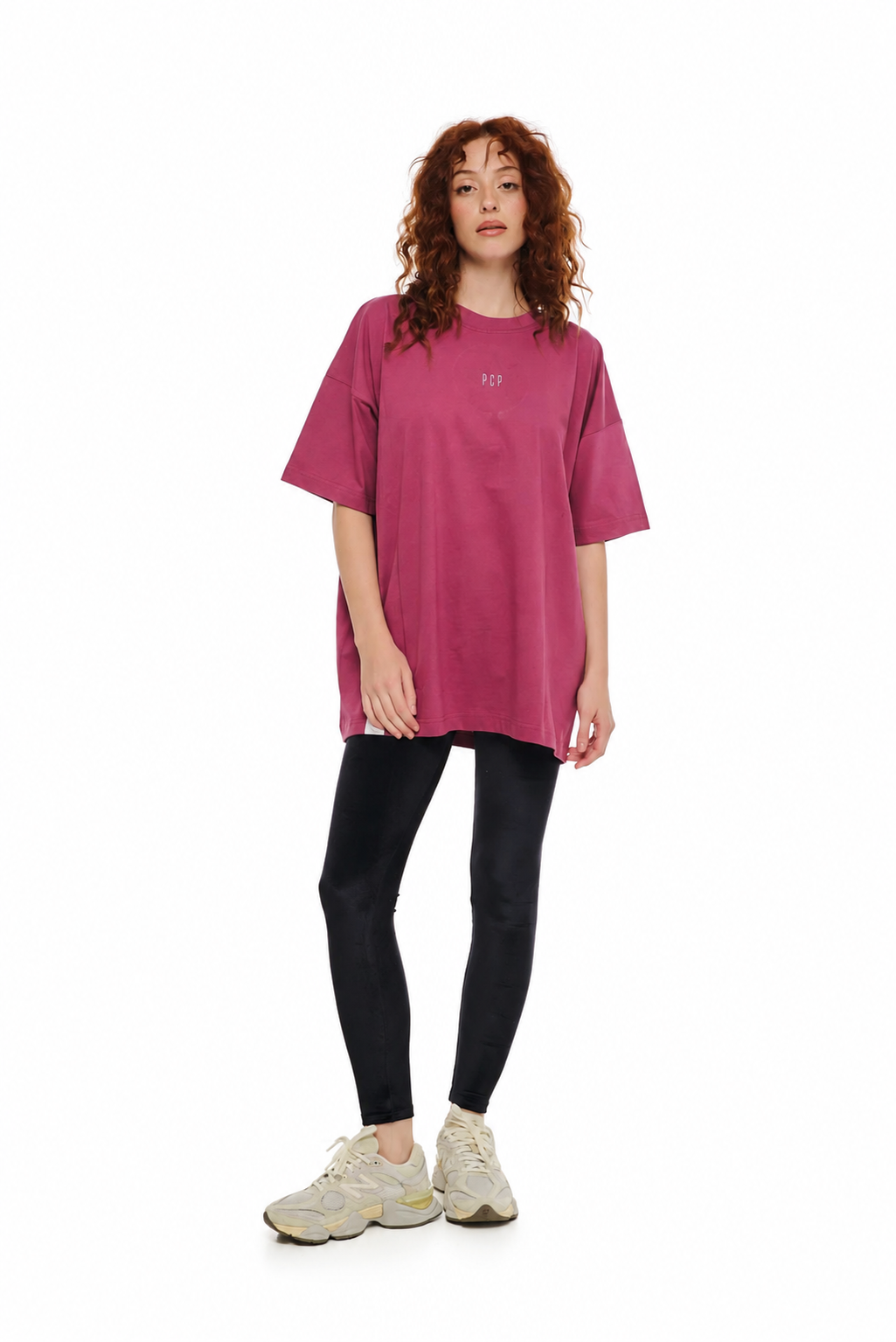 A woman modeling an oversized short-sleeve t-shirt in a dusty rose pink color, paired with black leggings and beige sneakers, against a plain white background.