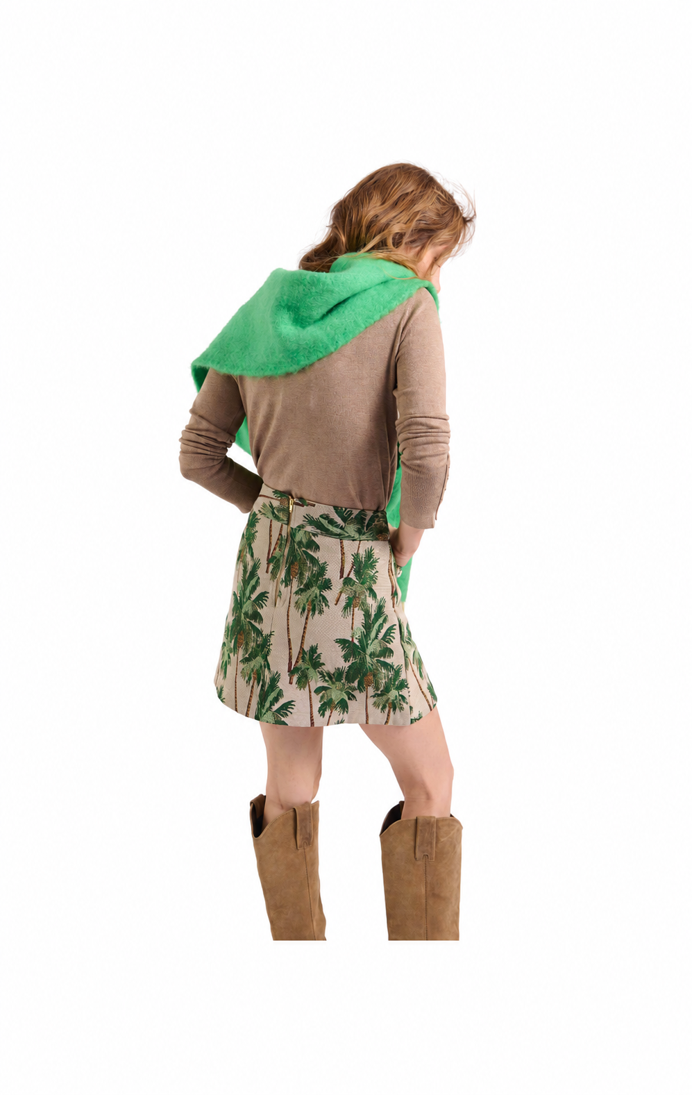 A woman shown from the back wearing a brown long-sleeve top, a beige mini skirt with a green palm tree print, and tan cowboy boots, with a bright green towel draped over her shoulders, against a plain white background.