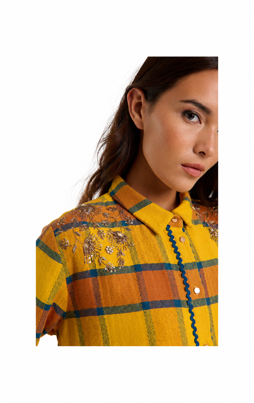 A close-up of a woman wearing a yellow plaid button-up shirt with orange and blue stripes, floral embroidery on the chest, and a blue scalloped trim along the placket, set against a plain white background.