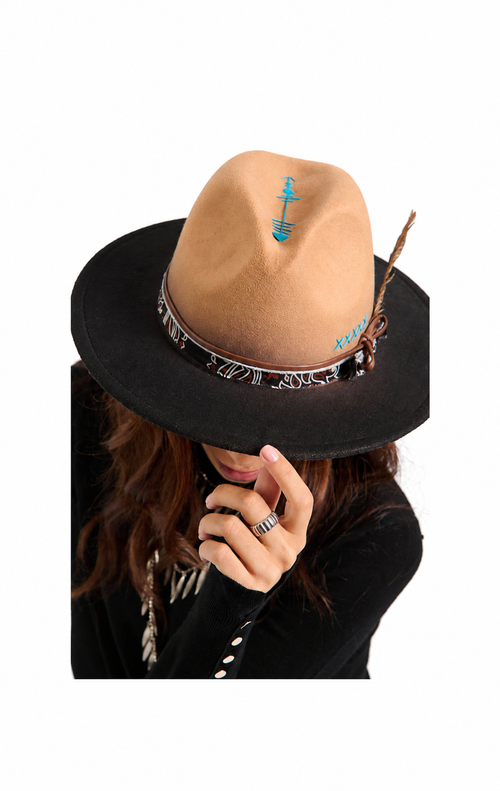 A woman wearing a tan felt fedora hat with a black brim, turquoise arrow decoration, and a feather accent, paired with a black top against a white background.