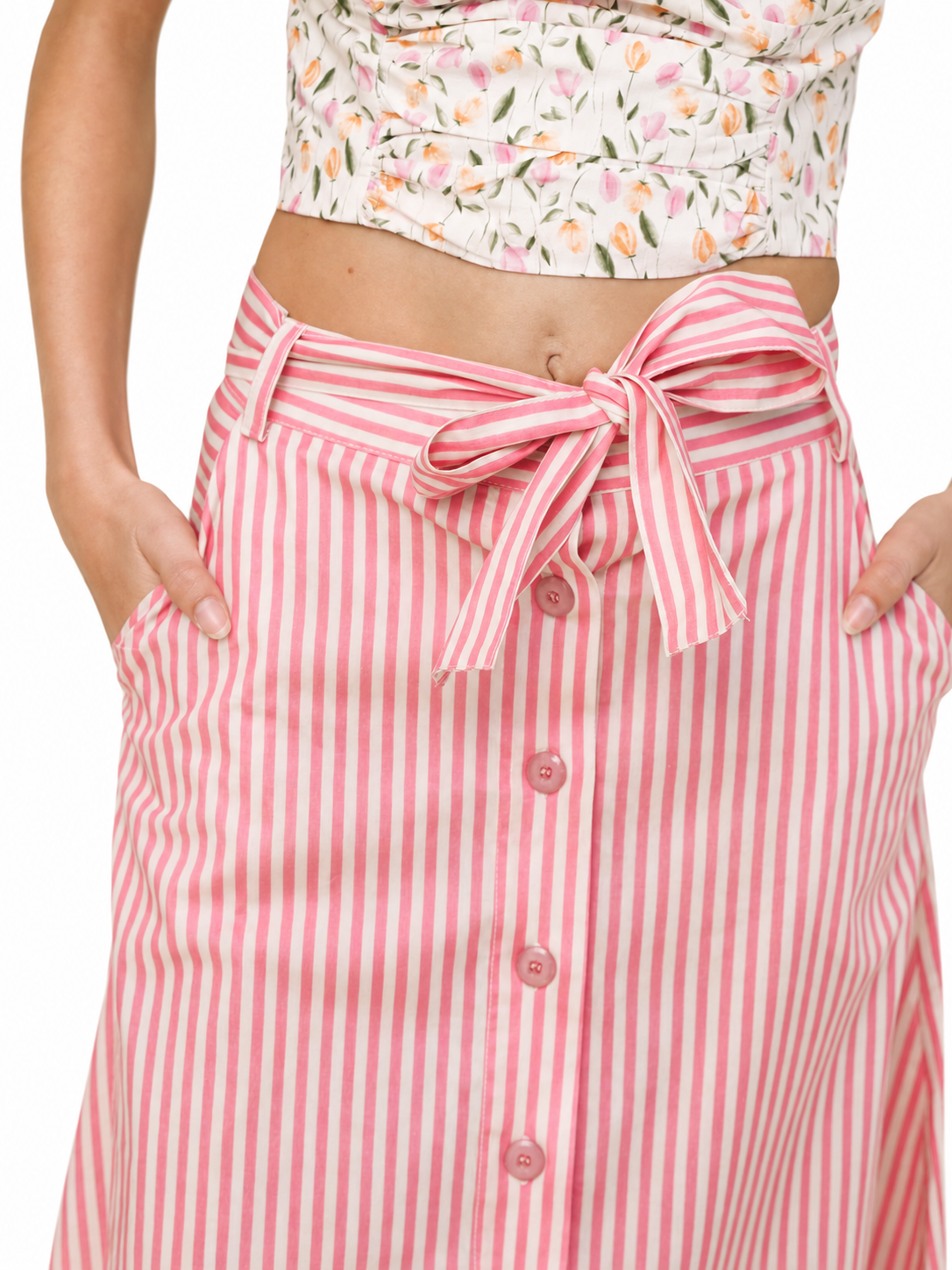 A close-up of a model wearing a pink and white vertically striped button-front skirt with a matching fabric tie belt at the waist. The model is also wearing a floral print crop top, shown from the waist down against a plain white background.