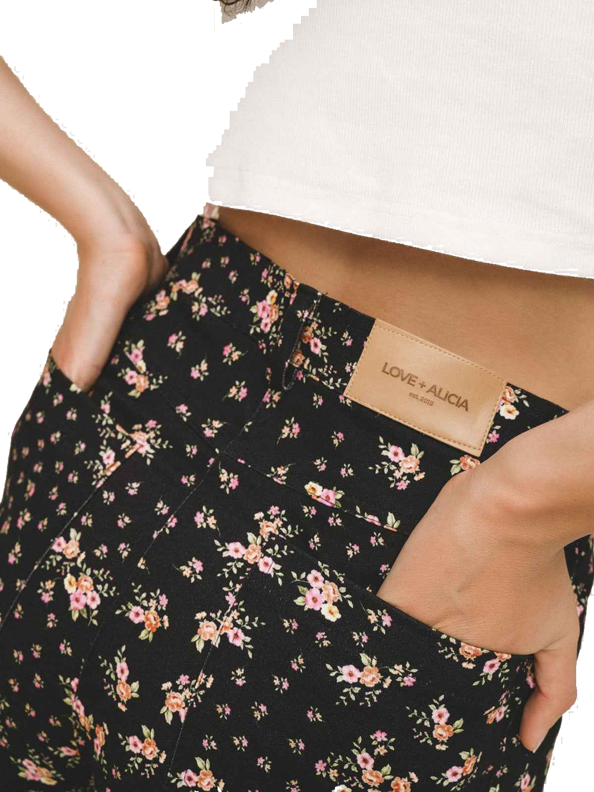 Close-up of a person wearing black floral pants with pink and yellow flowers, a white ribbed crop top, and hands in the pockets. A tan leather brand patch on the waistband reads LOVE+ALICIA.