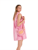 A woman wearing a pink sleeveless towel dress, holding a pink circular crossbody bag with a yellow eye design, against a solid white background.