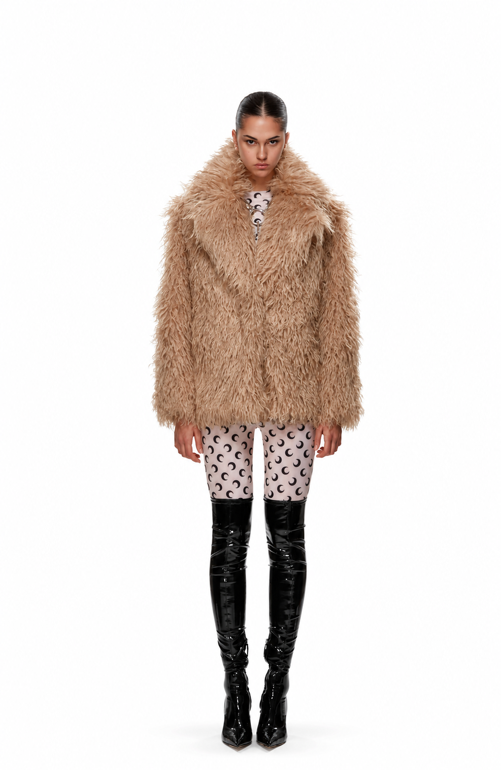 A model stands against a white background wearing a voluminous, shaggy beige faux-fur coat over a patterned top and black patent leather over-the-knee boots.