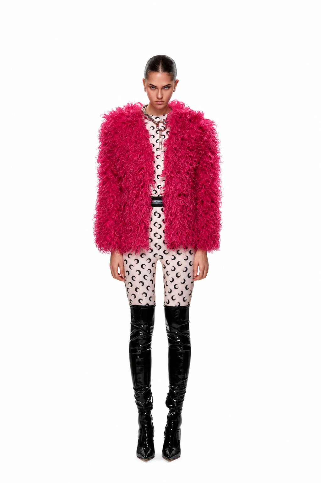 A model stands against a white background wearing a vibrant hot pink shaggy faux-fur jacket over a cream and black patterned shirt and shorts, paired with black patent leather knee-high boots.