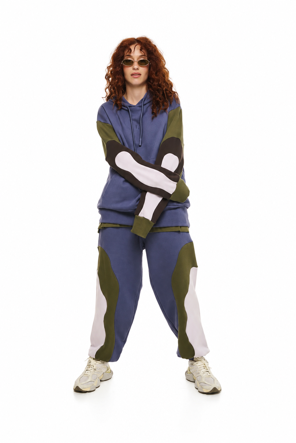 A model with curly red hair and sunglasses wearing a matching color-blocked hoodie and jogger pants set. The outfit features a slate blue base with olive green and off-white color-blocked panels, paired with off-white sneakers, against a solid white background.