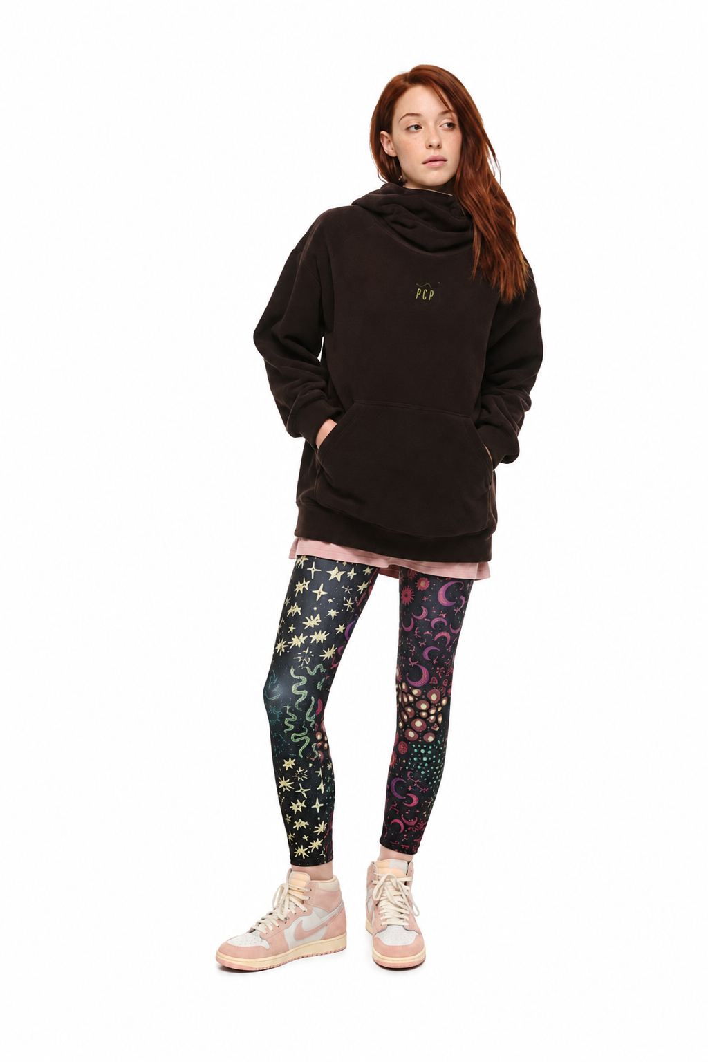 A model stands against a white background wearing a dark brown oversized hoodie, black leggings with colorful celestial prints, and light pink sneakers.