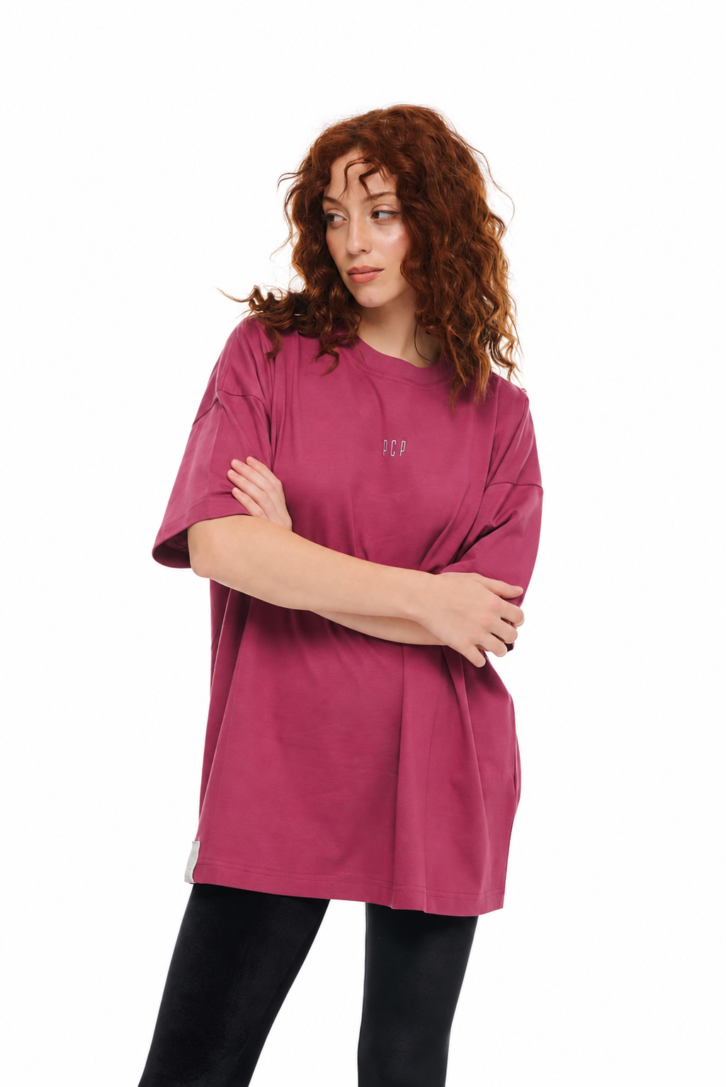 A woman with curly red hair wearing an oversized magenta-pink t-shirt with a small silver 'PCP' logo on the chest, paired with black leggings, against a plain white background.