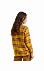 A woman shown from the back wearing a mustard yellow plaid flannel shirt with orange and blue stripes, paired with brown pants, against a solid white background.