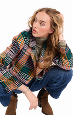 A woman crouching in a studio setting, wearing a multicolor plaid and check patterned jacket over a dark olive turtleneck, blue jeans, and brown suede boots.