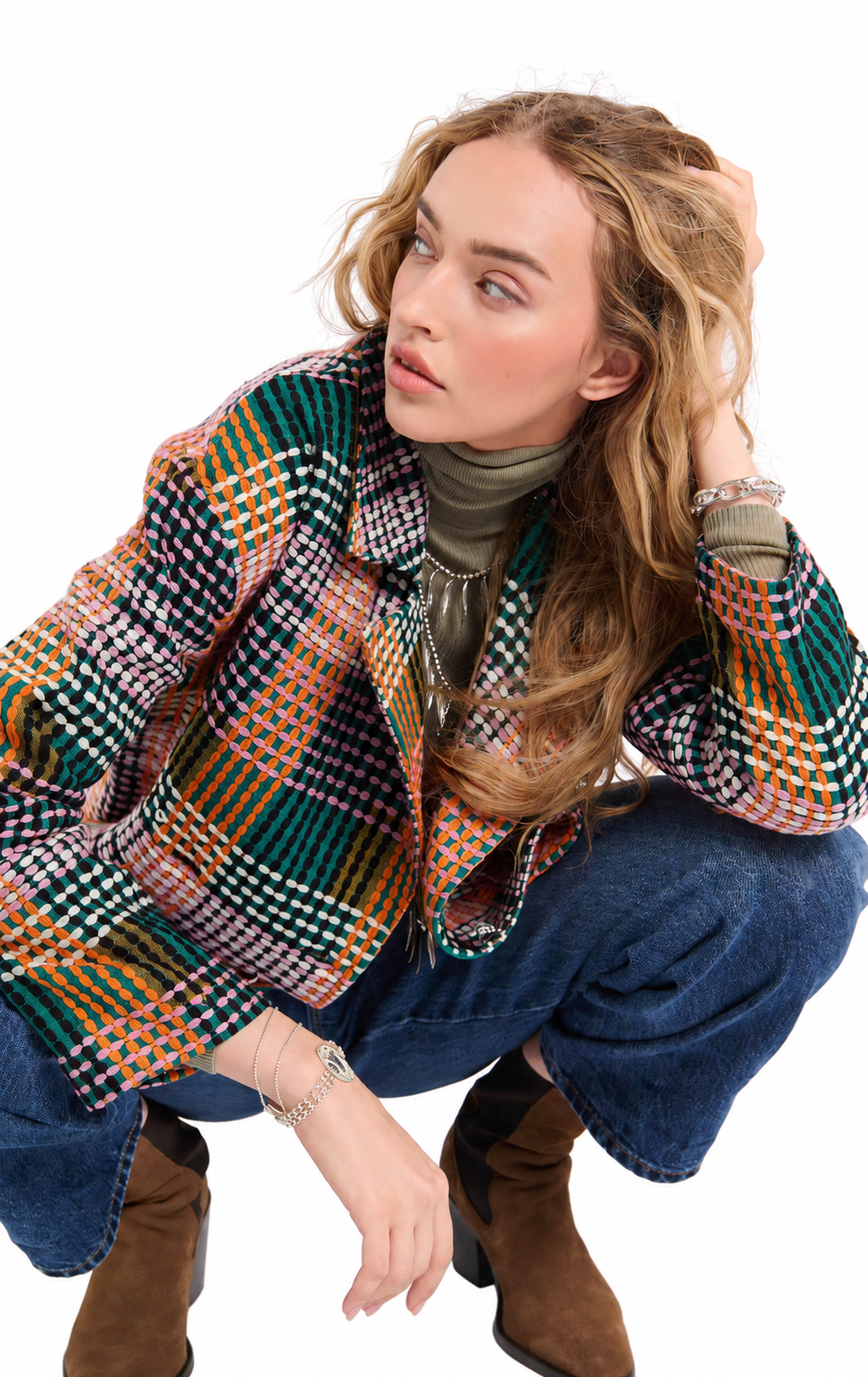 A woman crouching in a studio setting, wearing a multicolor plaid and check patterned jacket over a dark olive turtleneck, blue jeans, and brown suede boots.