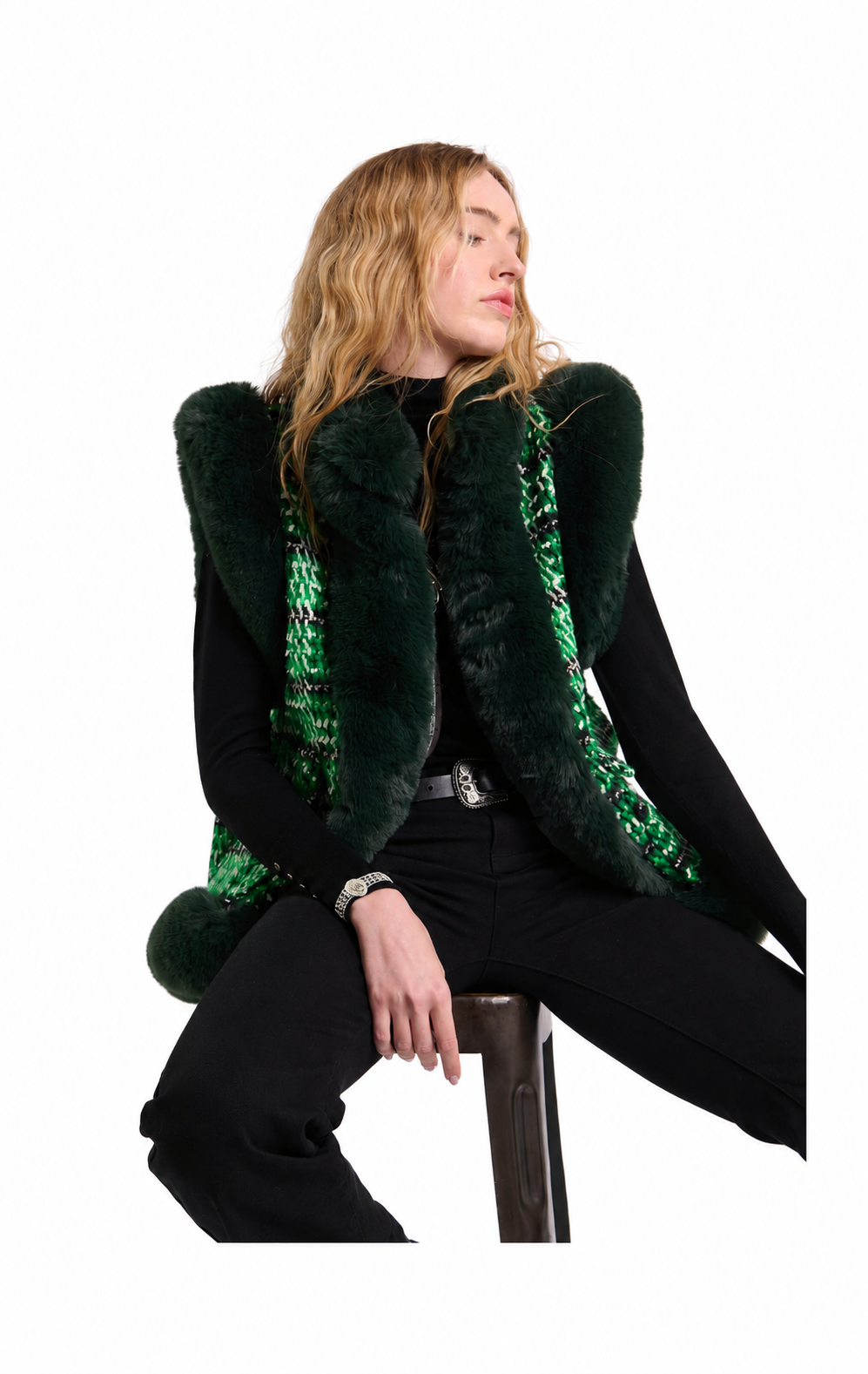 A woman seated on a stool wearing a dark green fur vest with bright green sequined trim over a black outfit, photographed against a plain white studio background.