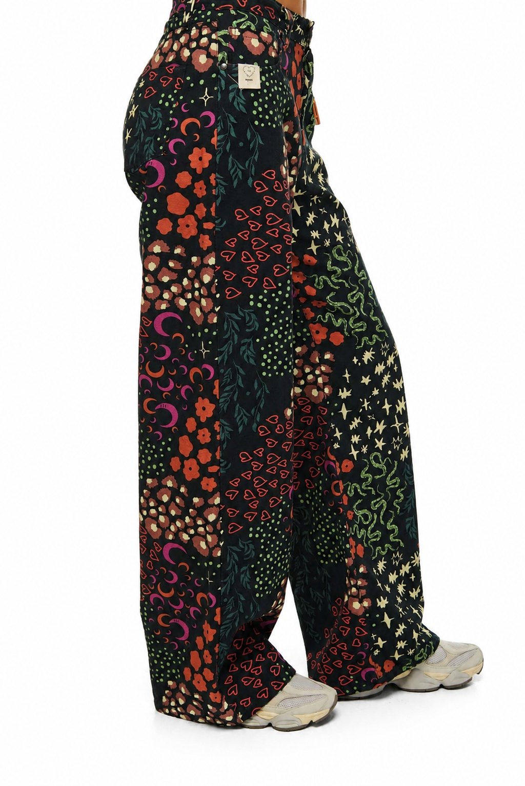 A person shown from the back wearing wide-leg patchwork pants with a multicolor floral and abstract print on a black base, paired with light gray sneakers, against a plain white background.