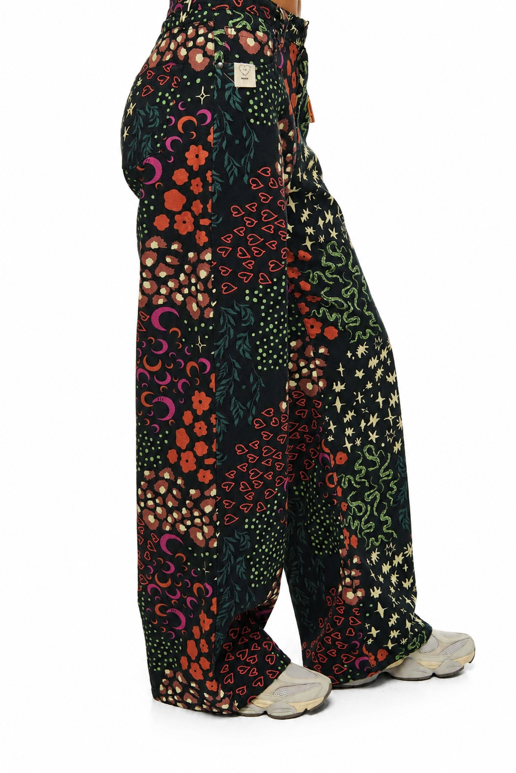 A person shown from the back wearing wide-leg patchwork pants with a multicolor floral and abstract print on a black base, paired with light gray sneakers, against a plain white background.
