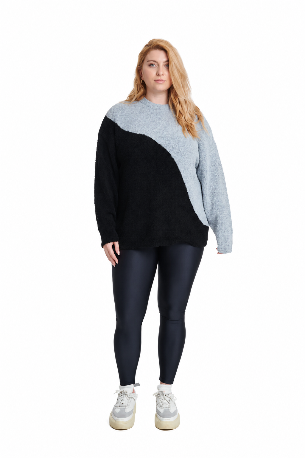 A woman modeling a color-block knit sweater with a light gray upper yoke and sleeves, and a black lower body, paired with dark leggings and white sneakers on a white background.