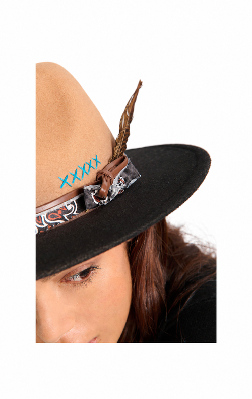 Close-up of a person wearing a black wide-brim hat with a tan crown, a brown feather, a patterned band, and blue 'XXXXX' text on the side.