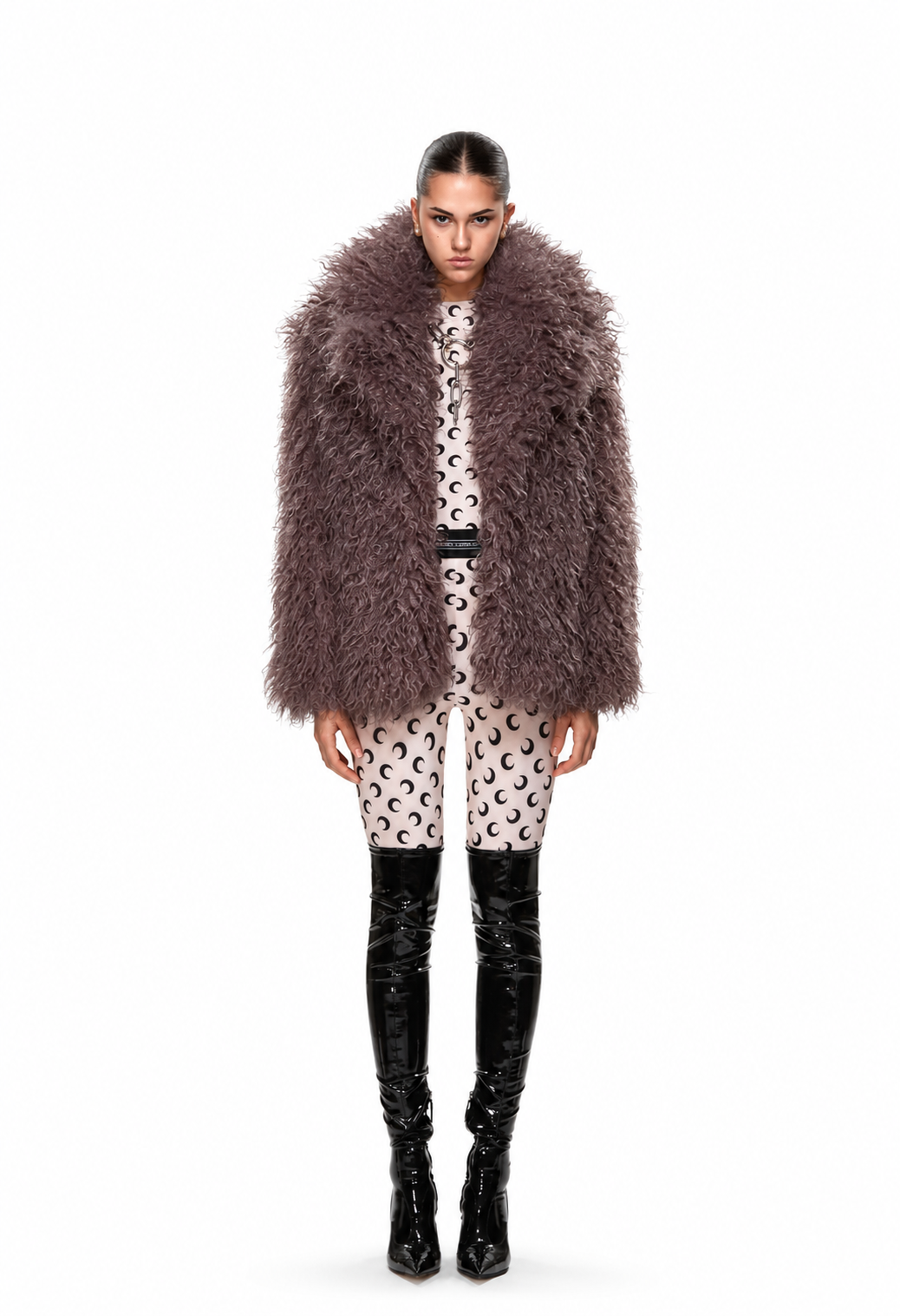 A model stands against a white studio background wearing a voluminous shaggy faux-fur jacket in taupe-brown, a beige and black patterned top and shorts, and black knee-high boots.