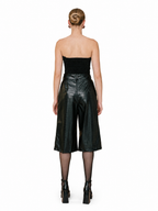 A model shown from the back wearing a black strapless bandeau top and shiny black faux-leather cropped wide-leg pants, paired with sheer black tights and black platform ankle boots, against a plain white background.