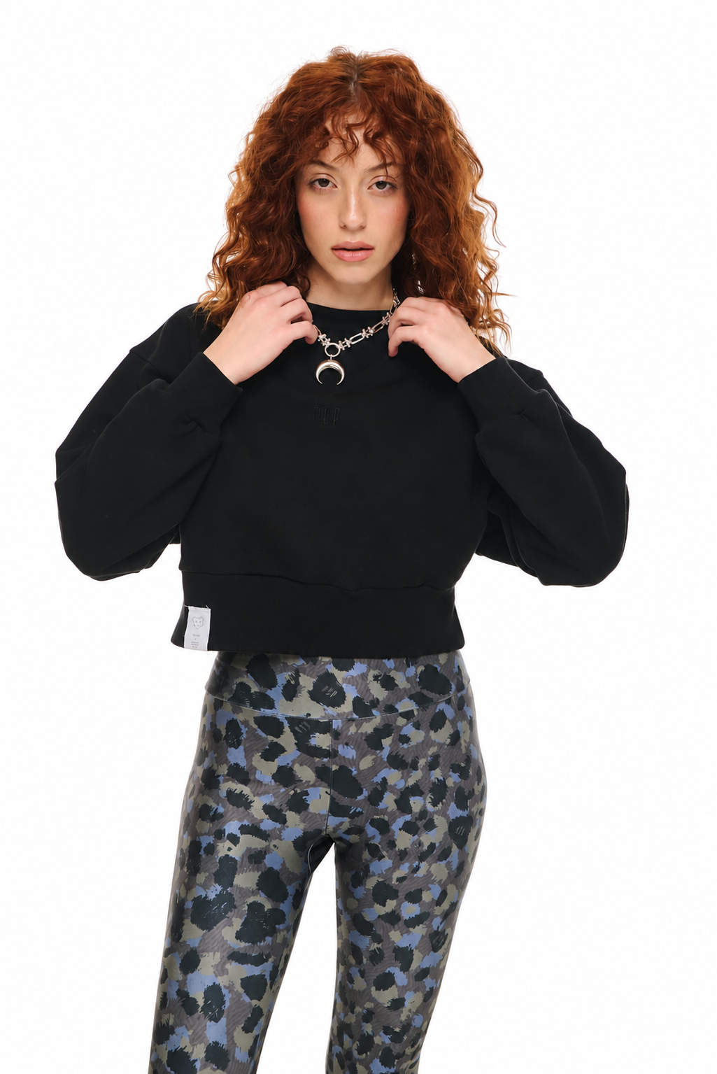 A model with curly red hair wearing a black cropped sweatshirt and blue-gray camouflage patterned leggings, posing against a solid white background.