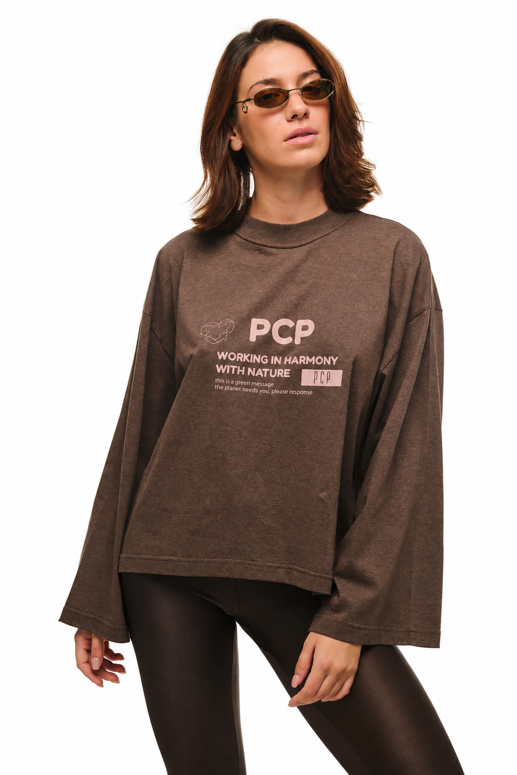 A woman modeling an oversized brown long-sleeve sweatshirt with a light pink graphic print on the front that reads 'PCP WORKING IN HARMONY WITH NATURE'. She is wearing dark brown leggings and sunglasses against a plain white background.