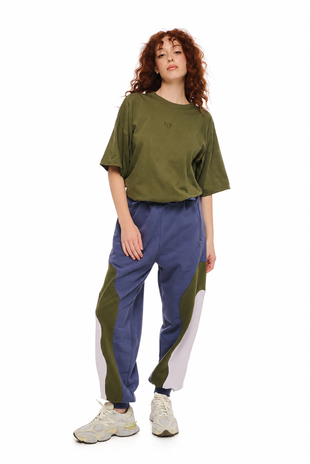 A model with curly red hair stands against a white background wearing a loose olive green t-shirt, colorblock sweatpants in green, blue, and white, and beige sneakers.