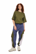 A model with curly red hair stands against a white background wearing a loose olive green t-shirt, colorblock sweatpants in green, blue, and white, and beige sneakers.