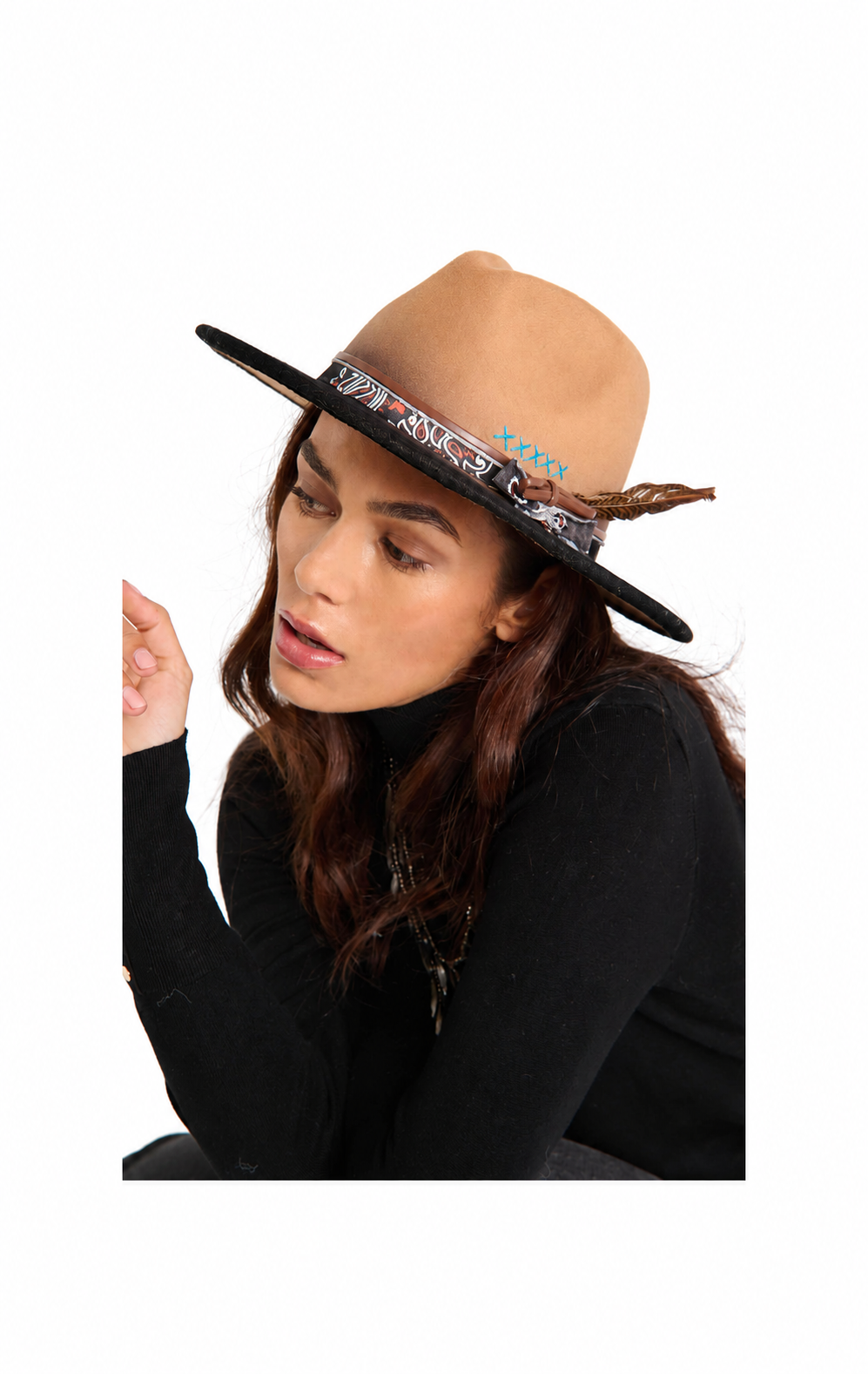 A woman wearing a tan wide-brim fedora hat with a patterned black band and a feather accent, paired with a black turtleneck, set against a plain white background.