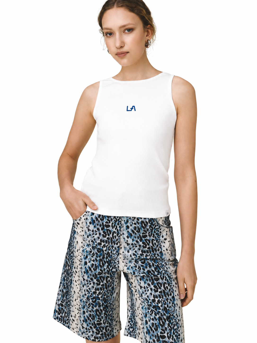 A woman modeling a white sleeveless tank top with a small blue 'LA' logo on the chest, paired with blue, black, and white leopard-print shorts, against a plain white background.
