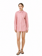A woman modeling a pink and white vertically striped button-down shirt dress, paired with black strappy flat sandals, against a solid white background.