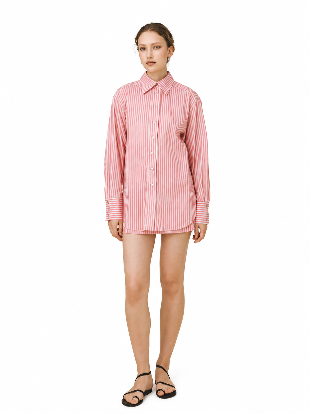 A woman modeling a pink and white vertically striped button-down shirt dress, paired with black strappy flat sandals, against a solid white background.