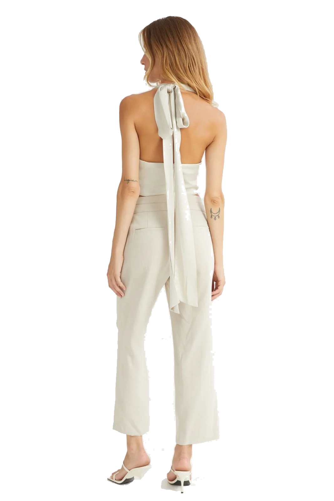 A woman shown from the back wearing a light cream halter jumpsuit with a large tie detail at the back of the neck and matching cropped trousers, paired with white heeled sandals against a plain white background.