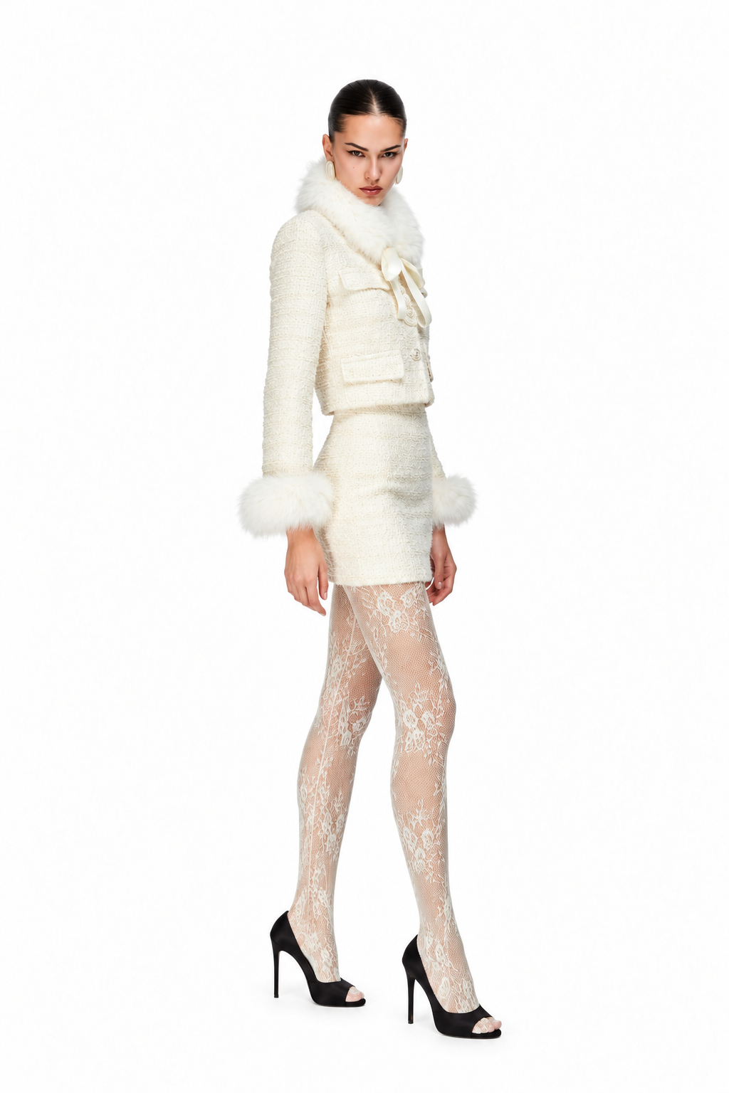 A model wearing a white textured jacket and matching skirt with faux fur trim, paired with white lace tights and black high heels, set against a plain white background.