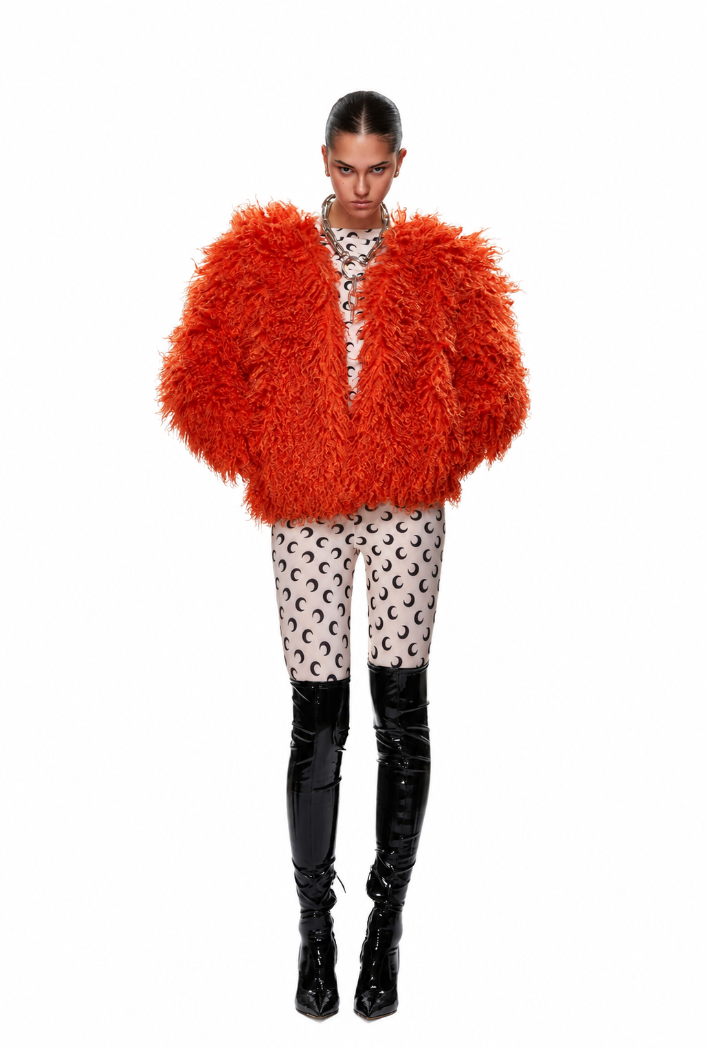 A model wearing a vibrant orange shaggy faux-fur jacket, patterned beige and black pants, and black patent leather boots, shot against a solid white background.
