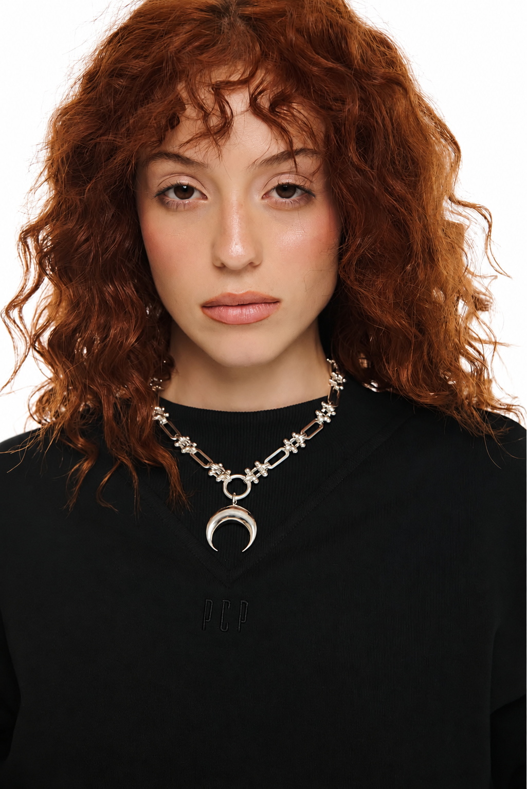 A close-up of a model with curly red hair wearing a silver-toned chain necklace with star and crescent moon charms, paired with a black top against a white background.