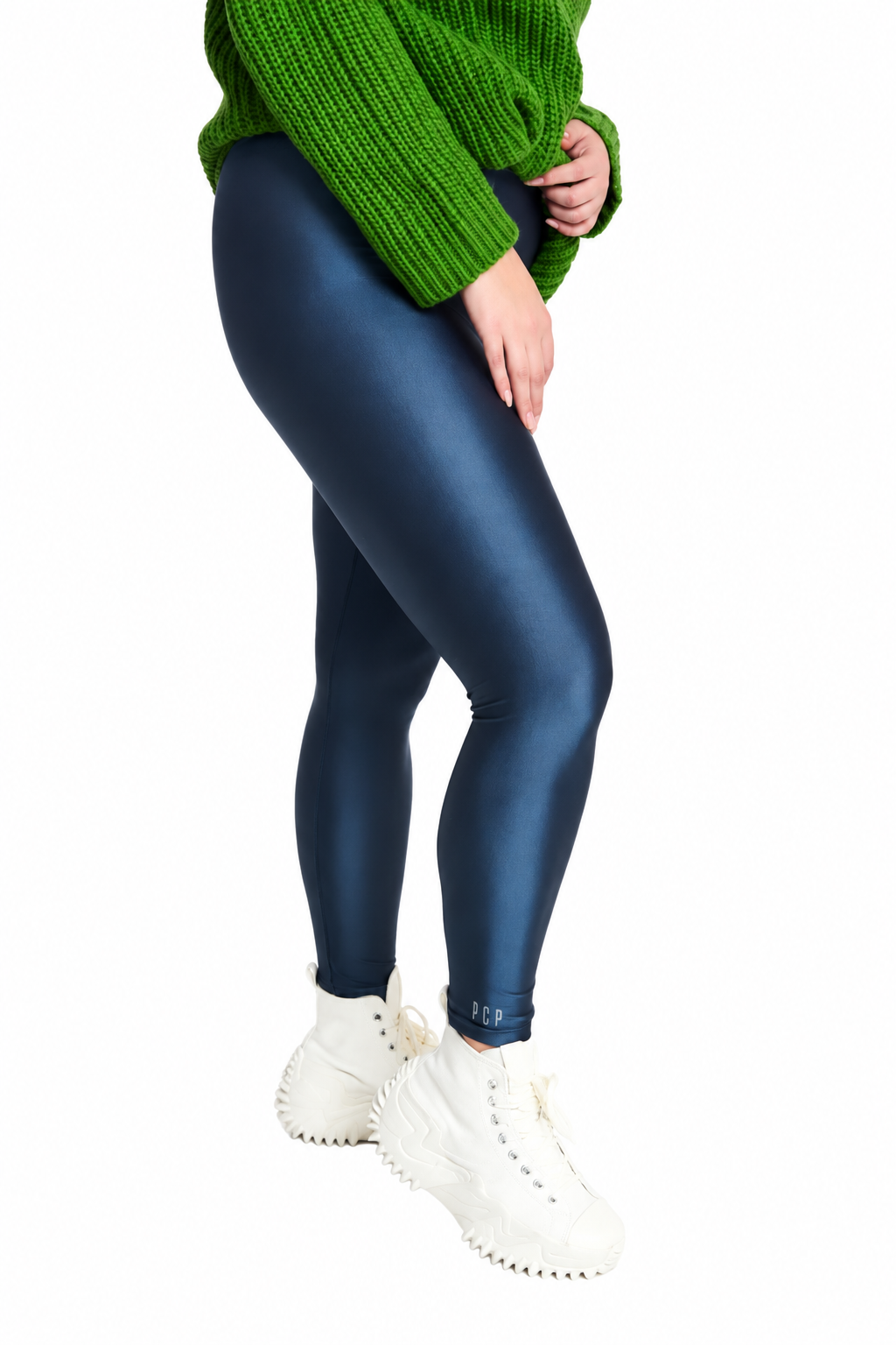A person wearing shiny dark blue leggings, a green knit sweater, and white sneakers, shown from the waist down against a plain white background.
