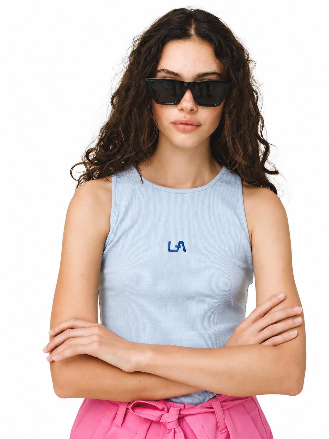 A woman with long curly brown hair wearing black sunglasses, a light blue sleeveless tank top with a blue 'LA' logo on the chest, and pink pants, posed against a solid white background.