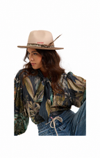 A woman wearing a beige wide-brim hat with a feather and a botanical print blouse tucked into blue jeans, photographed against a plain white background.