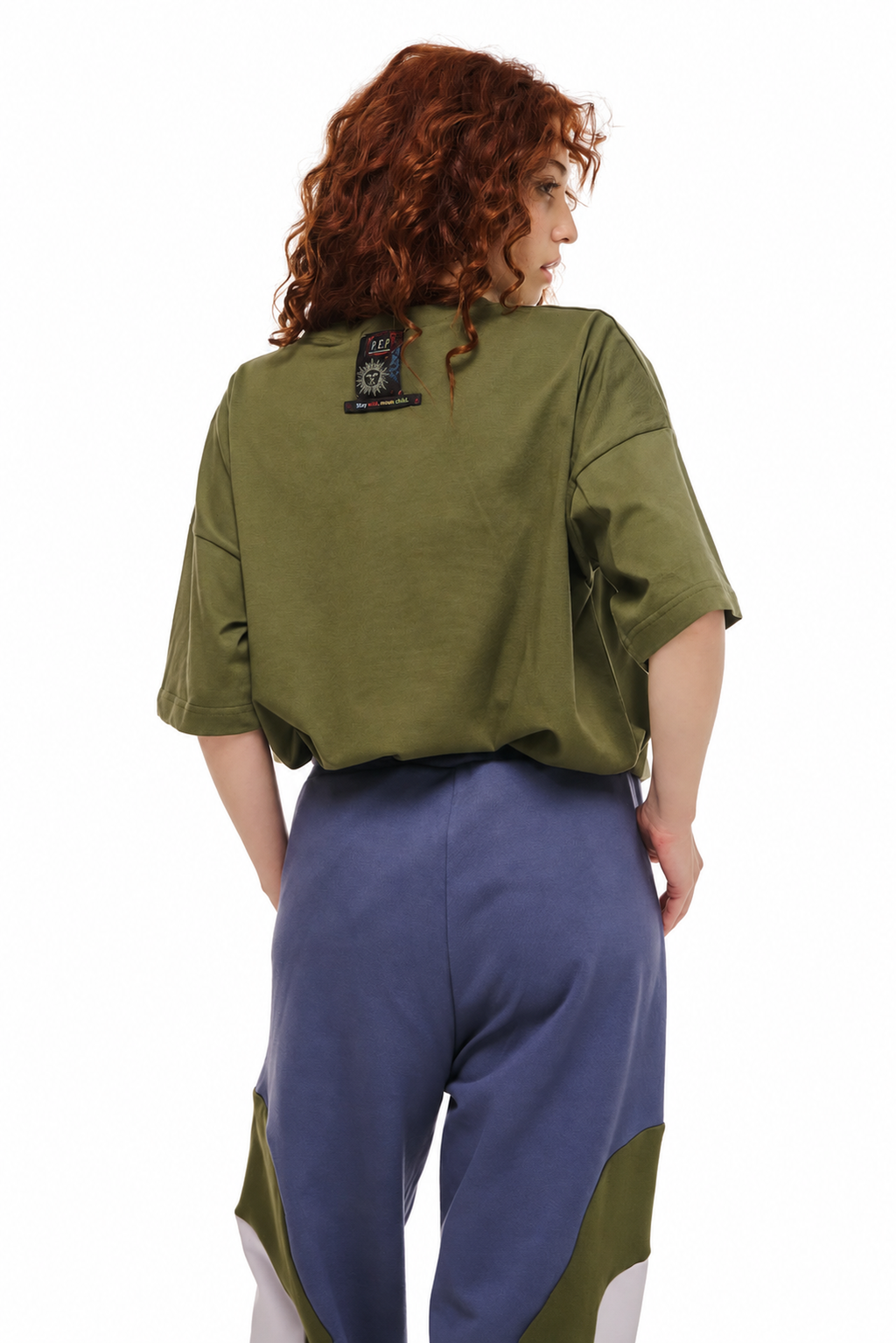 A model with curly red hair is shown from the back wearing an olive green cropped short-sleeve shirt and blue pants with olive green and white color-blocked details, against a plain white background.