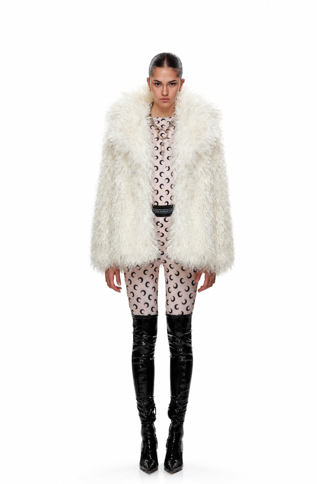 A model stands against a white studio background wearing a voluminous white faux-fur jacket over a pink and black patterned top and matching pants, paired with black patent leather over-the-knee boots.