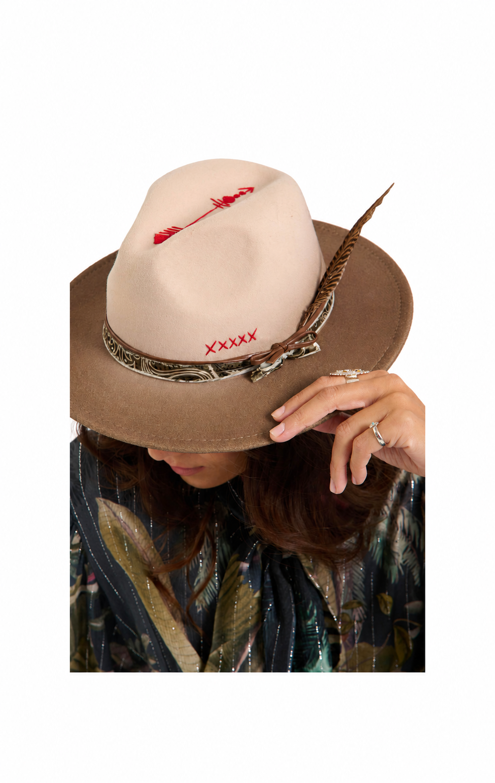 A woman wearing a beige and brown fedora hat with red embroidered details, a feather accent, and a patterned scarf, shown against a plain white background.