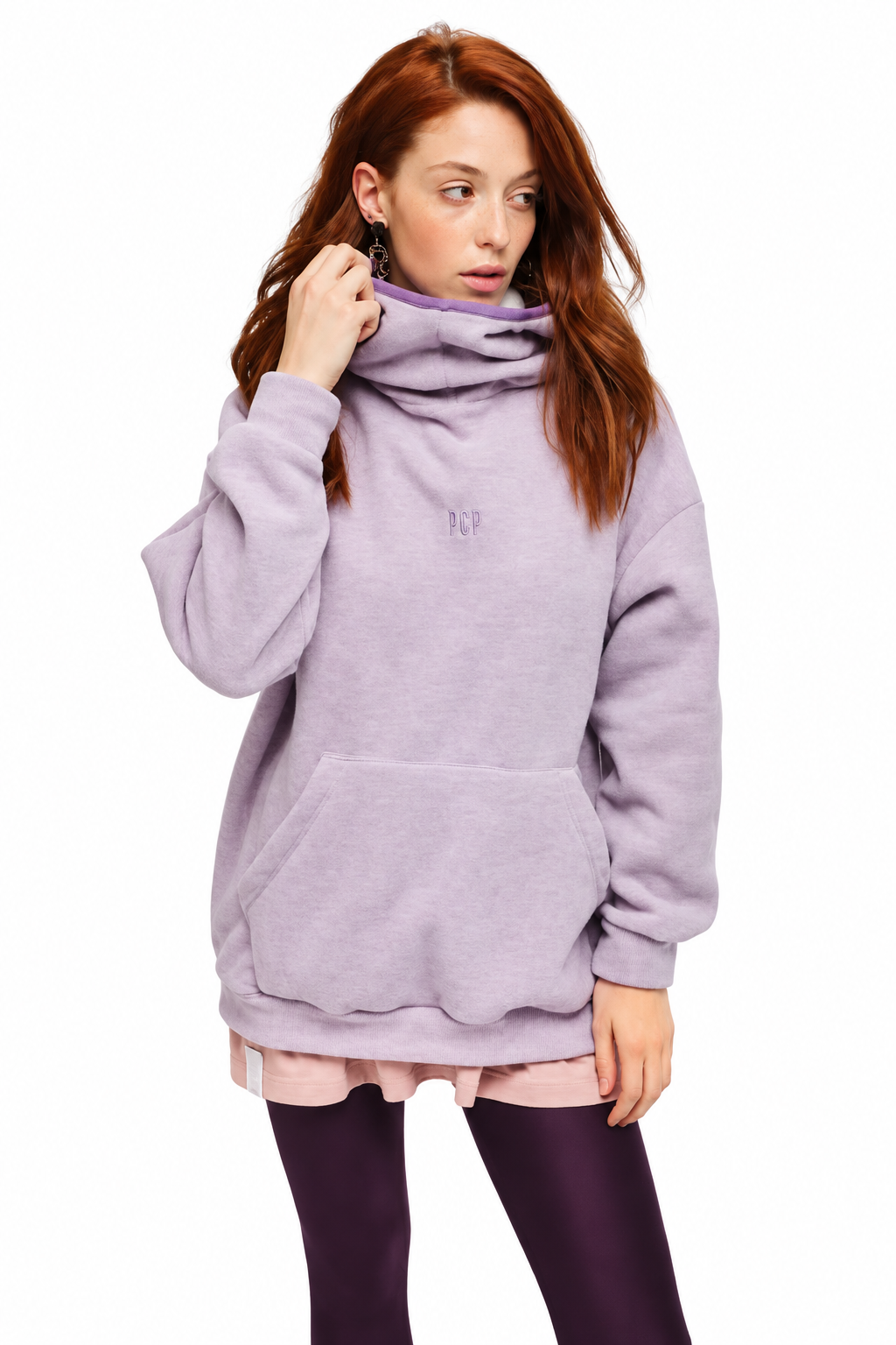 A woman with red hair wearing a lavender oversized cowl-neck hoodie, a light pink shirt underneath, and dark purple leggings, photographed against a solid white background.
