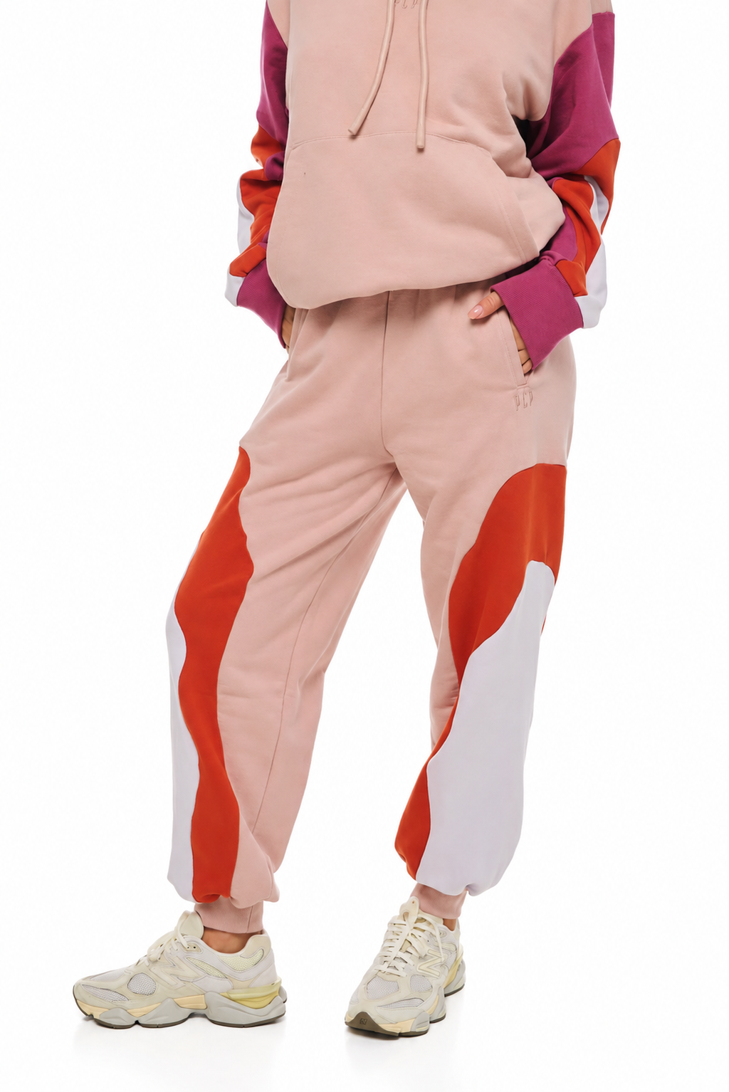 A person wearing a color-blocked hoodie and jogger sweatpants set in pastel pink, orange-red, and white, paired with chunky cream sneakers, against a plain white background.
