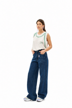 A woman modeling a white sleeveless tank top with green trim and a green 'Polo' logo, paired with high-waisted, wide-leg blue jeans and white sneakers, against a solid white background.