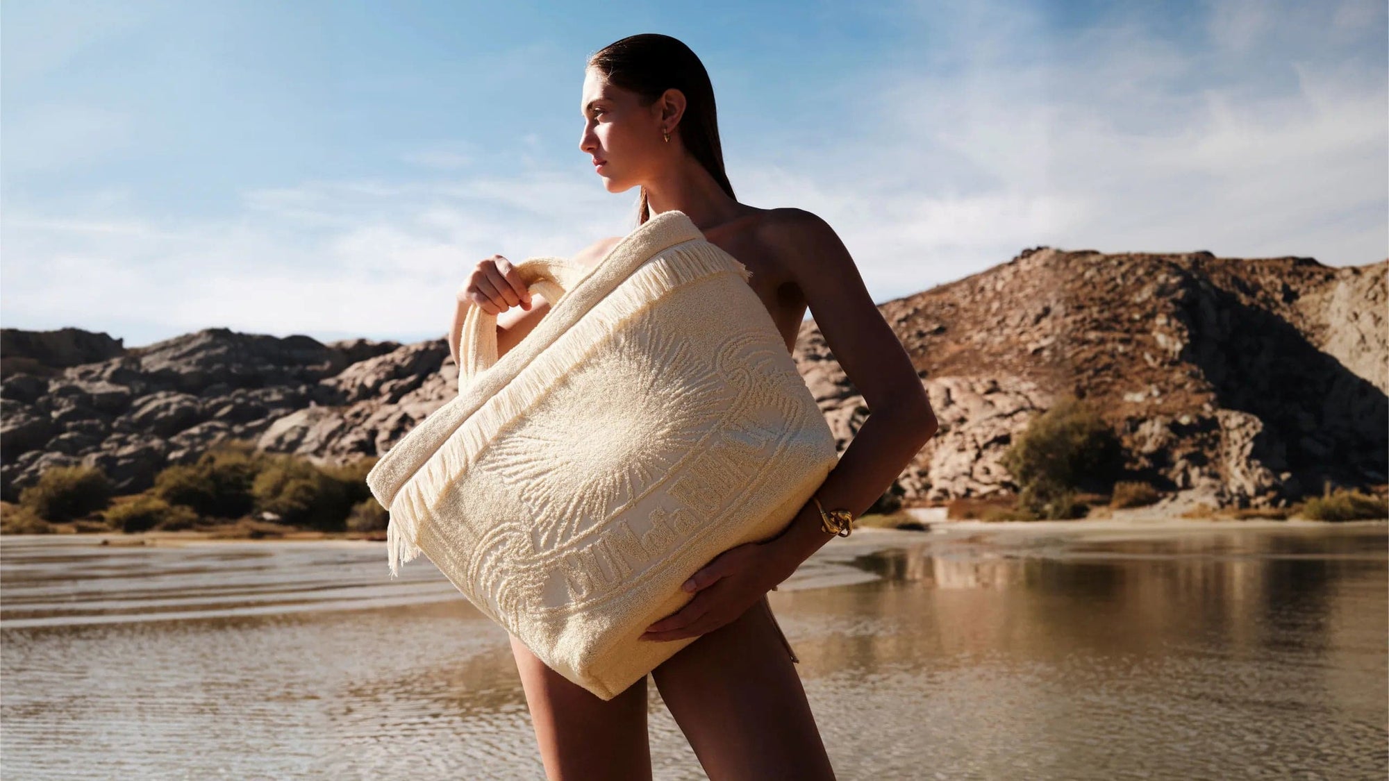 Woman wrapped in a towel by a body of water with mountains in the background