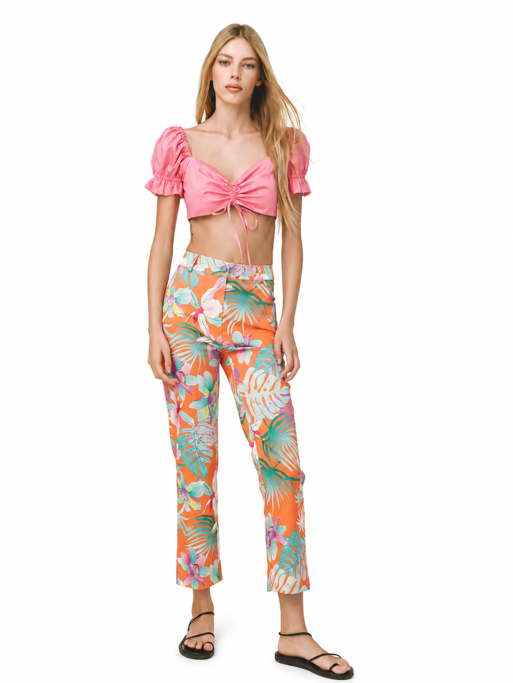 A model wearing a two-piece outfit consisting of a pink puff-sleeve crop top with a front tie detail and high-waisted cropped pants featuring a vibrant orange, green, and white tropical leaf print, set against a plain white background.