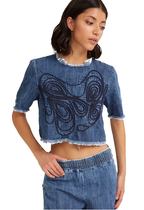 A woman wearing a cropped blue denim top with a large dark abstract embroidered design on the front, paired with matching denim shorts, against a plain white background.