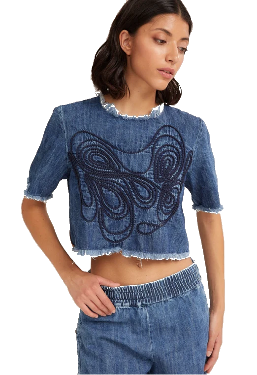 A woman wearing a cropped blue denim top with a large dark abstract embroidered design on the front, paired with matching denim shorts, against a plain white background.