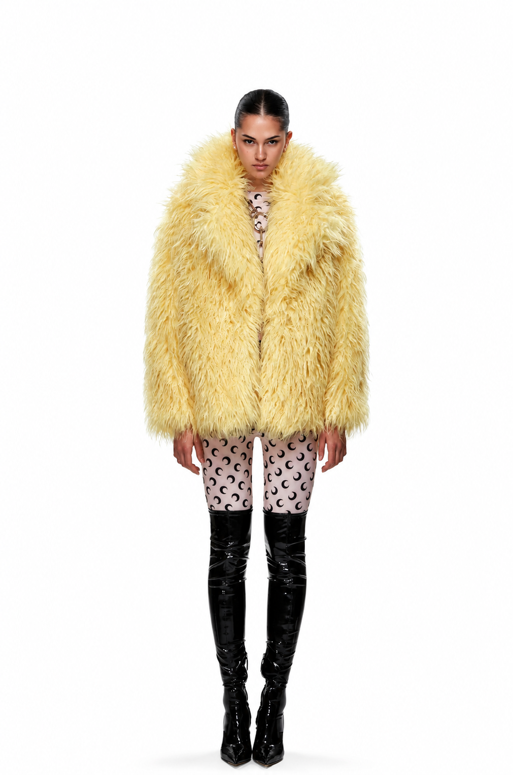 A model stands against a white studio background wearing a voluminous pale yellow faux-fur coat over a patterned top and black thigh-high glossy boots.